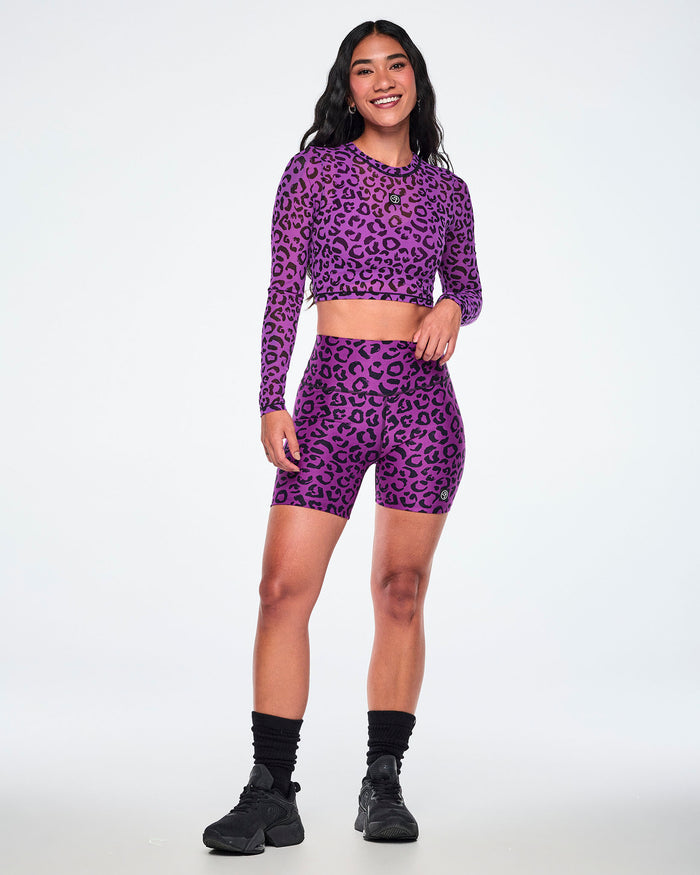 Full view of a female model wearing a matching purple and black leopard-print activewear set, featuring a long-sleeve mesh crop top and high-waisted biker shorts, paired with black socks and shoes.