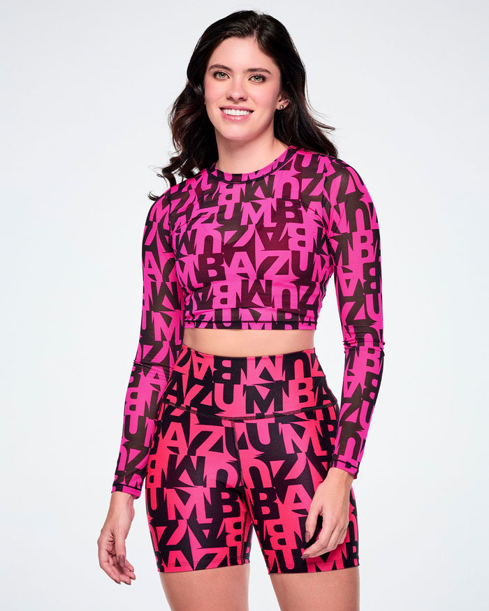 A female model wearing a matching two-piece activewear set featuring a long-sleeve mesh crop top and high-waisted biker shorts, both printed with a bold abstract pink and black ZUMBA lettering, on a white background.