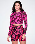 A female model wearing a matching two-piece activewear set featuring a long-sleeve mesh crop top and high-waisted biker shorts, both printed with a bold abstract pink and black ZUMBA lettering, on a white background.
