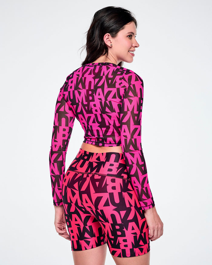 Back view of a female model wearing a matching two-piece activewear set featuring a long-sleeve mesh crop top and high-waisted biker shorts, both printed with a bold abstract pink and black ZUMBA lettering, on a white background.