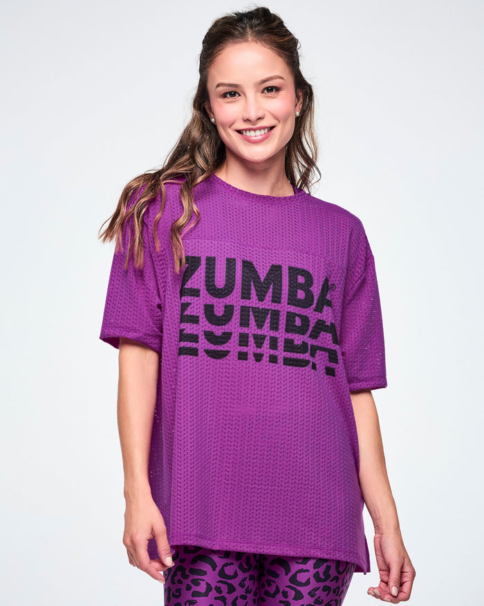 Female model in a purple oversized mesh boxy top with a large black stacked “ZUMBA” logo across the chest, paired with purple leopard-print biker shorts against a white background.