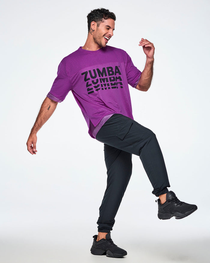 A male model wearing a purple mesh top with a black 'ZUMBA' graphic stacked on the front, paired with black pants and black sneakers on a white background.