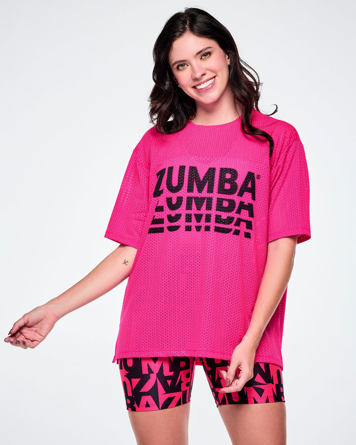 Female model wearing a hot pink oversized mesh boxy top with a large black “ZUMBA” stacked logo across the chest, paired with black and pink biker shorts featuring an all-over bold “ZUMBA” lettering pattern.