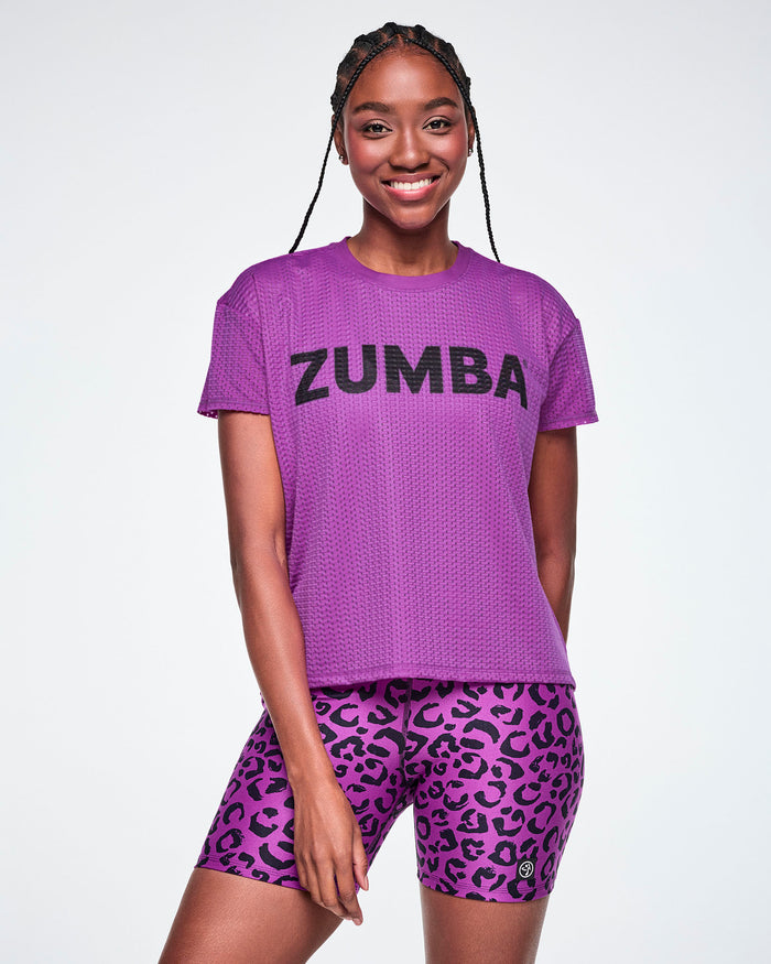 A female model wearing a purple Zumba short-sleeve mesh top with black 'ZUMBA' text across the chest, paired with purple leopard print biker shorts, against a white background.