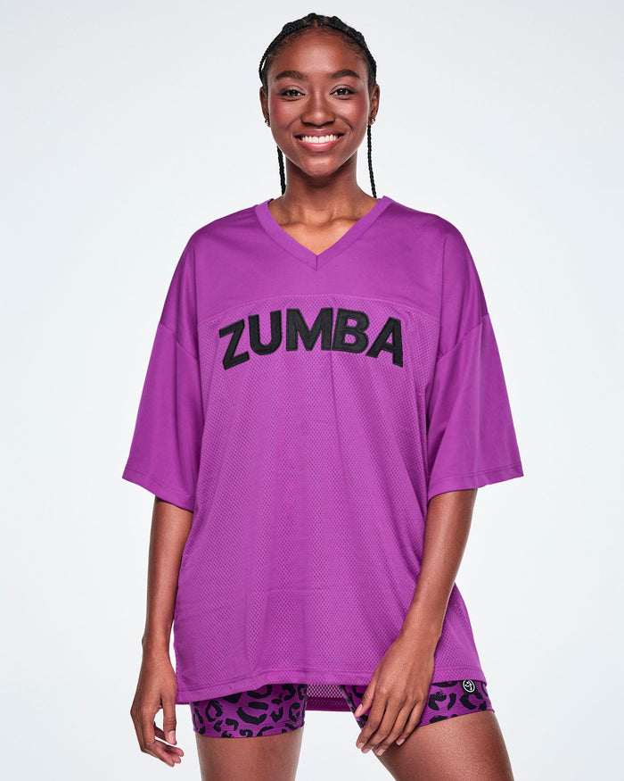 Female model wearing an oversized purple mesh V-neck top with “ZUMBA” printed in black across the chest, paired with purple leopard print high-waisted biker shorts.
