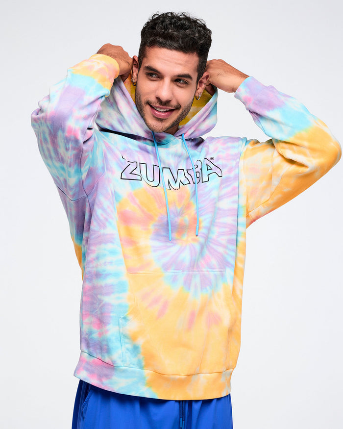Male model wearing a tie-dye Zumba hoodie grabbing the hoodie with the drawstrings hanging over the black outlined Zumba on the chest.