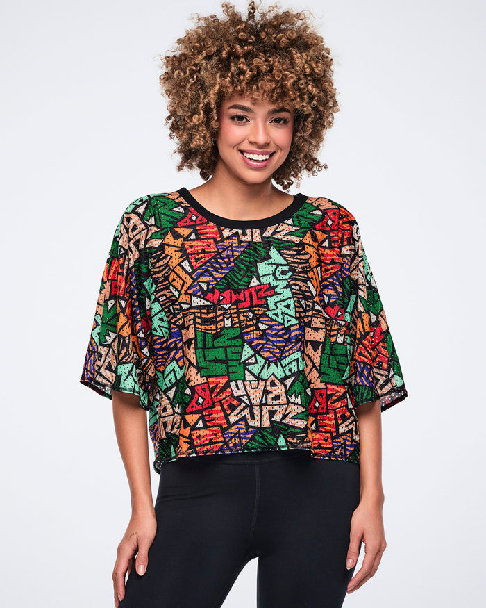 Multicolor abstract-print cropped mesh top with a black crew neckline and loose elbow-length sleeves. Female model wears the top over black high-waisted leggings, shown in a front-facing studio pose.