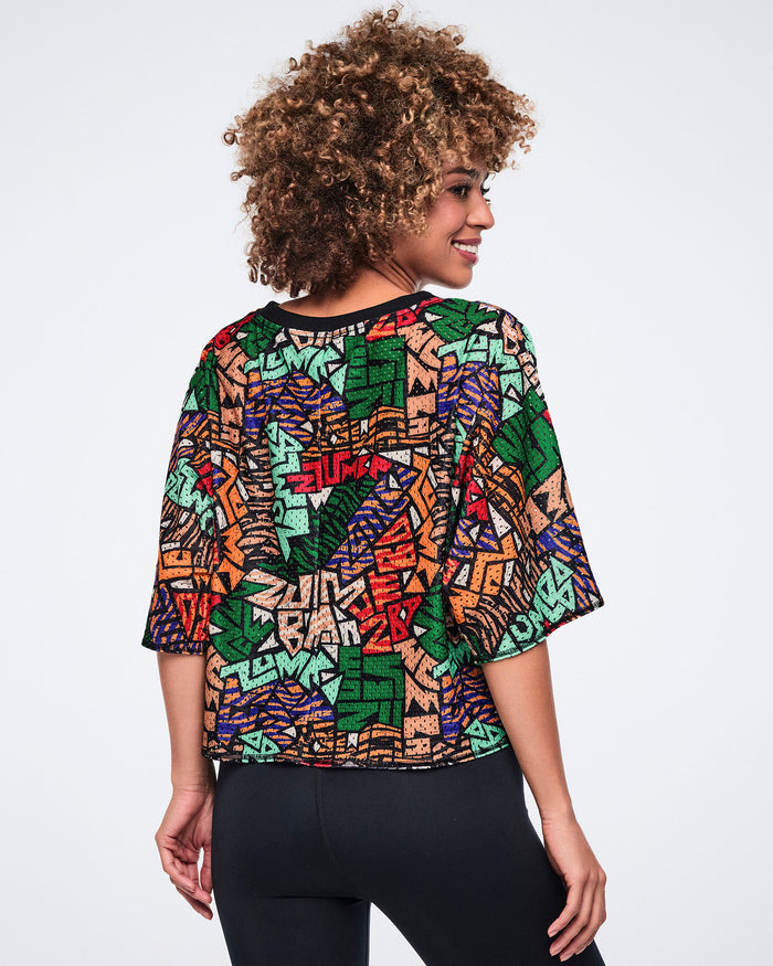 Back view of the multicolor abstract-print cropped mesh top with a black crew neckline and loose elbow-length sleeves. Female model wears the top with black high-waisted leggings, shown turned over her shoulder.
