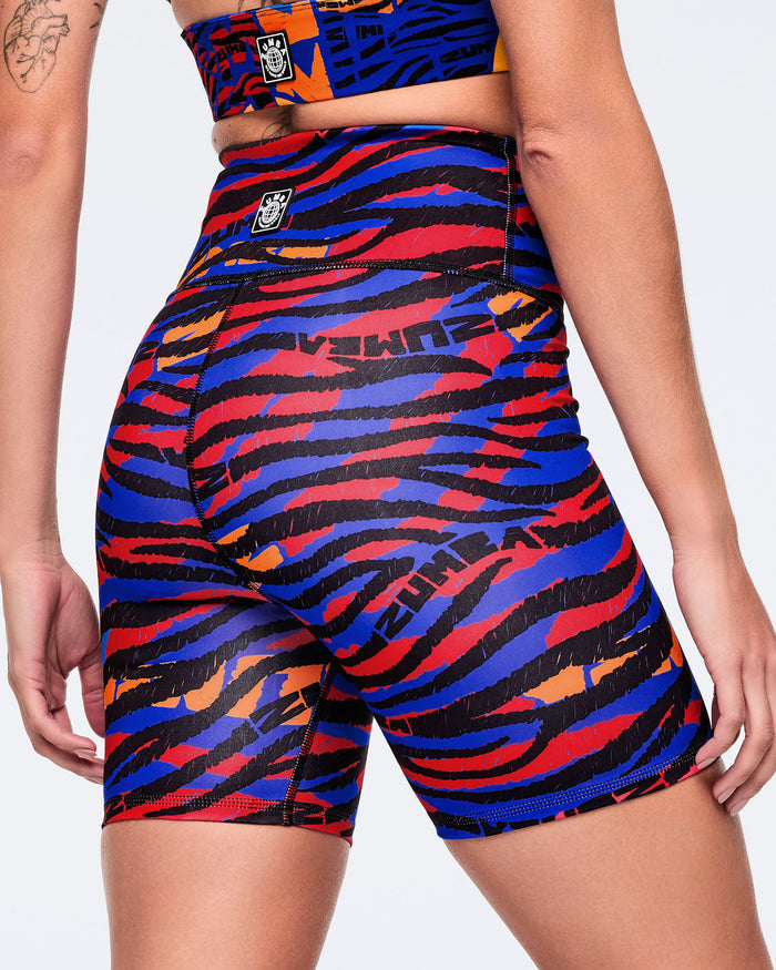 Female model wearing high-waisted biker shorts in a vivid blue, red, black, and orange abstract zebra-print design, shown from the back with contour seams and a smooth, body-hugging fit.