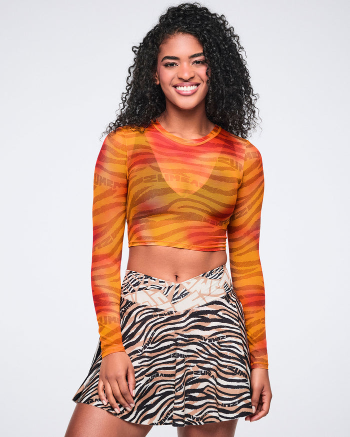 Orange and red zebra-print sheer long-sleeve crop top with a crew neckline and cropped fitted silhouette. Female model wears the top with a beige, black, and cream zebra-print flared mini skirt.