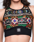 Close-up of a multicolor geometric-print longline sports bra with a high neckline, black trim, and a wide black under-band with a centered Zumba logo patch. Female model wears the bra in a cropped front view.