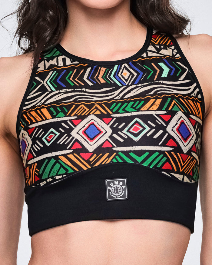 Close-up of a multicolor geometric-print longline sports bra with a high neckline, black trim, and a wide black under-band with a centered Zumba logo patch. Female model wears the bra in a cropped front view.