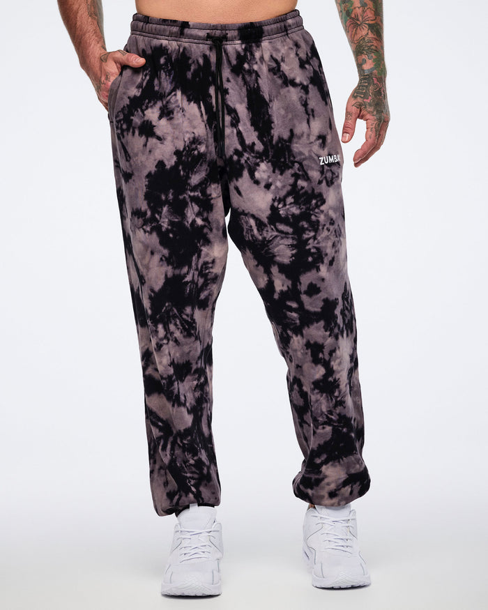 Black and tan tie-dye sweatpants with a small white Zumba logo on the front. Male model wears the relaxed-fit sweatpants with a black drawstring waistband, side pockets, tapered legs, and white low-top sneakers.