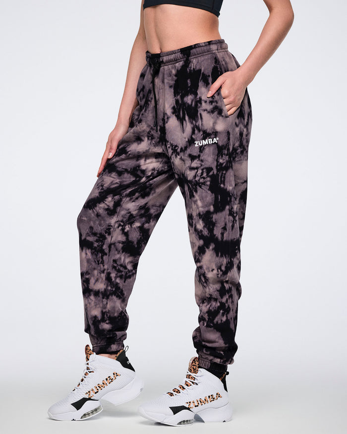 Black and tan tie-dye sweatpants with a small white Zumba logo on the front. Female model wears the relaxed-fit sweatpants with a black drawstring waistband, side pockets, cuffed ankles, a black sports bra, and white high-top sneakers with black and tan accents.