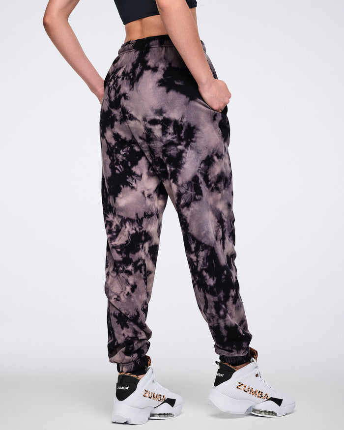 Black and tan tie-dye sweatpants shown from the back. Female model wears the relaxed-fit sweatpants with an elastic waistband, cuffed ankles, a black sports bra, and white high-top sneakers with black and tan accents.
