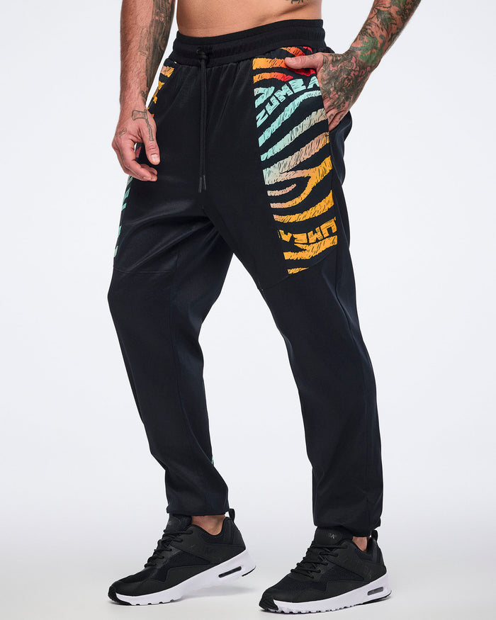 Male model wearing black jogger pants with multicolor zebra-print paneling on the upper legs, a drawstring waistband, tapered legs, and black sneakers with white soles.