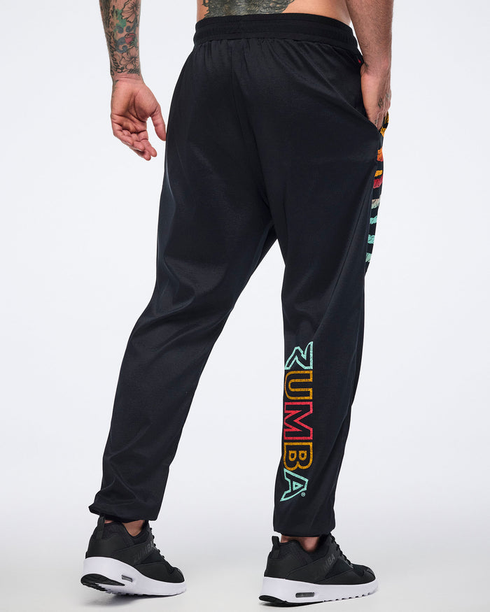Male model wearing black jogger pants shown from the back with a relaxed fit, multicolor Zumba logo on the lower leg, tapered cuffs, and black sneakers with white soles.