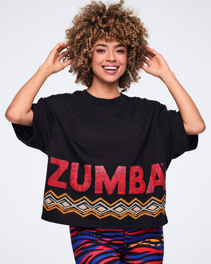 Female model wearing a black oversized cropped T-shirt with bold red Zumba lettering and an orange, white, and black geometric border print along the hem, paired with multicolor zebra-print leggings.