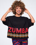 Female model wearing a black oversized cropped T-shirt with bold red Zumba lettering and an orange, white, and black geometric border print along the hem, paired with multicolor zebra-print leggings.