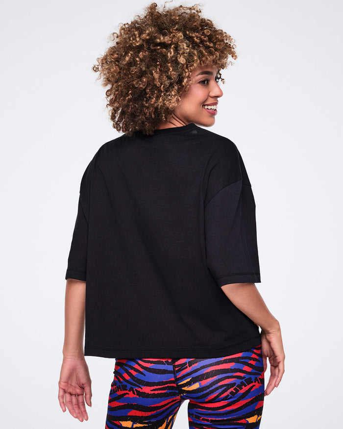 Female model wearing a black oversized cropped T-shirt shown from the back with short sleeves, a loose fit, and multicolor zebra-print leggings.