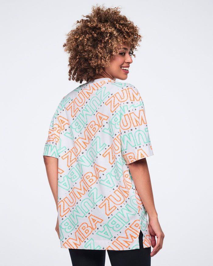Back view of a white short-sleeve Zumba oversized T-shirt with a crew neckline and allover mint and orange diagonal logo print. Female model wears the tunic-length tee with black leggings.