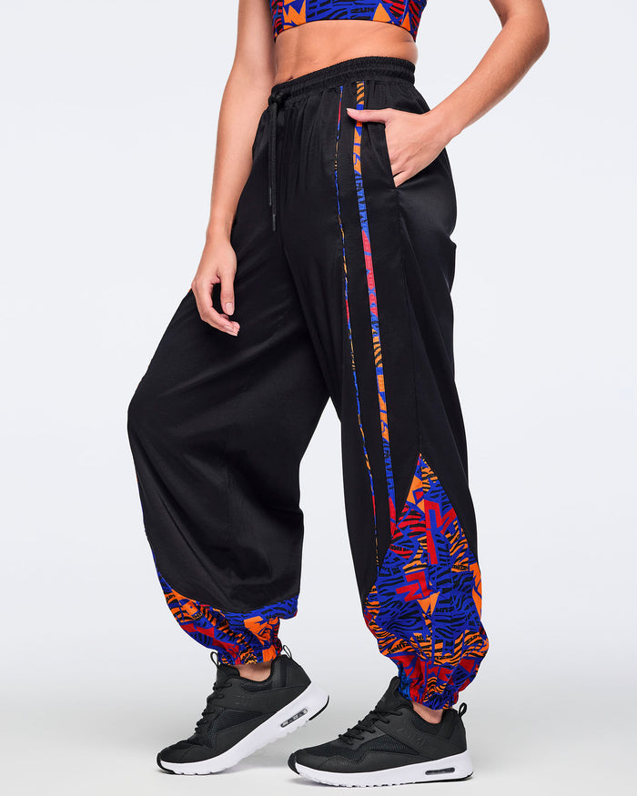 Side view of black loose-fit dance pants with an elastic drawstring waistband, side pockets, and blue, orange, and red abstract print trim and lower-leg panels. Female model wears the pants with a matching blue and orange crop top and black athletic sneakers.