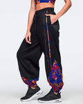 Side view of black loose-fit dance pants with an elastic drawstring waistband, side pockets, and blue, orange, and red abstract print trim and lower-leg panels. Female model wears the pants with a matching blue and orange crop top and black athletic sneakers.