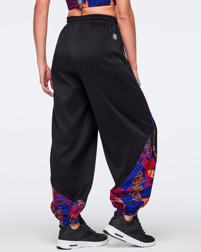 Back view of black loose-fit dance pants with a draped silhouette, elastic waistband, and blue, orange, and red abstract print panels at the lower legs. Female model wears the pants with a matching blue and orange crop top and black athletic sneakers.