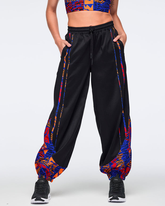 Front view of black loose-fit dance pants with an elastic drawstring waistband, side pockets, and blue, orange, and red abstract print trim and gathered cuffs. Female model wears the pants with a matching blue and orange crop top and black athletic sneakers.