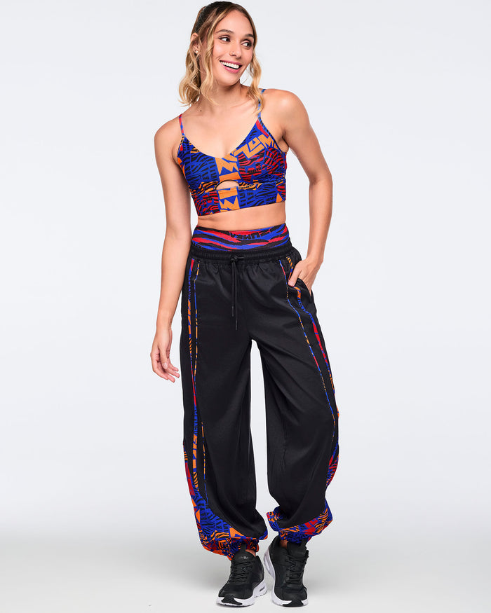 Black loose-fit dance pants with an elastic drawstring waistband, side pockets, and blue, orange, and red abstract print trim and gathered cuffs. Female model wears the pants with a matching blue and orange crop top and black athletic sneakers.