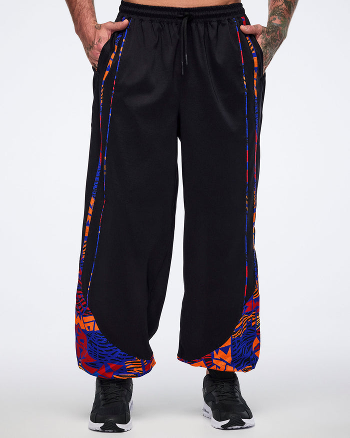 Front view of black loose-fit dance pants with an elastic drawstring waistband, side pockets, and blue, orange, and red abstract print panels at the lower legs. Male model wears the pants with black athletic sneakers.