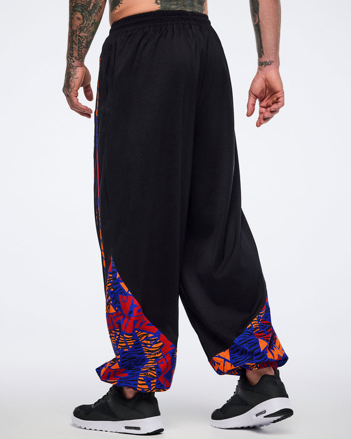 Back view of black loose-fit dance pants with a draped silhouette, elastic waistband, and blue, orange, and red abstract print panels at the lower legs. Male model wears the pants with black athletic sneakers.