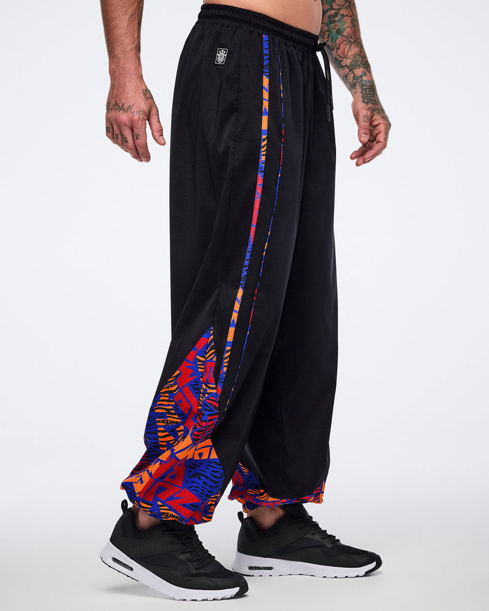 Side view of black loose-fit dance pants with an elastic drawstring waistband, side pockets, and blue, orange, and red abstract print trim and lower-leg panels. Male model wears the pants with black athletic sneakers.