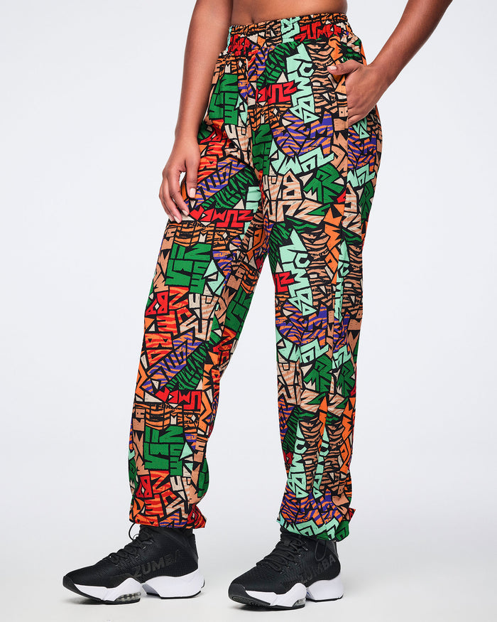 Side view of multicolor Zumba print track pants with an elastic waistband, relaxed straight-leg fit, side pockets, and adjustable ankle bungee cords. Female model wears the pants with black athletic sneakers.