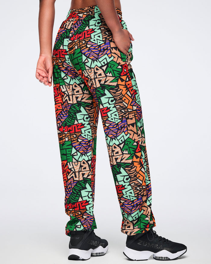 Back view of multicolor Zumba print track pants with an elastic waistband, relaxed fit, side pockets, and adjustable ankle bungee cords. Female model wears the pants with black athletic sneakers.