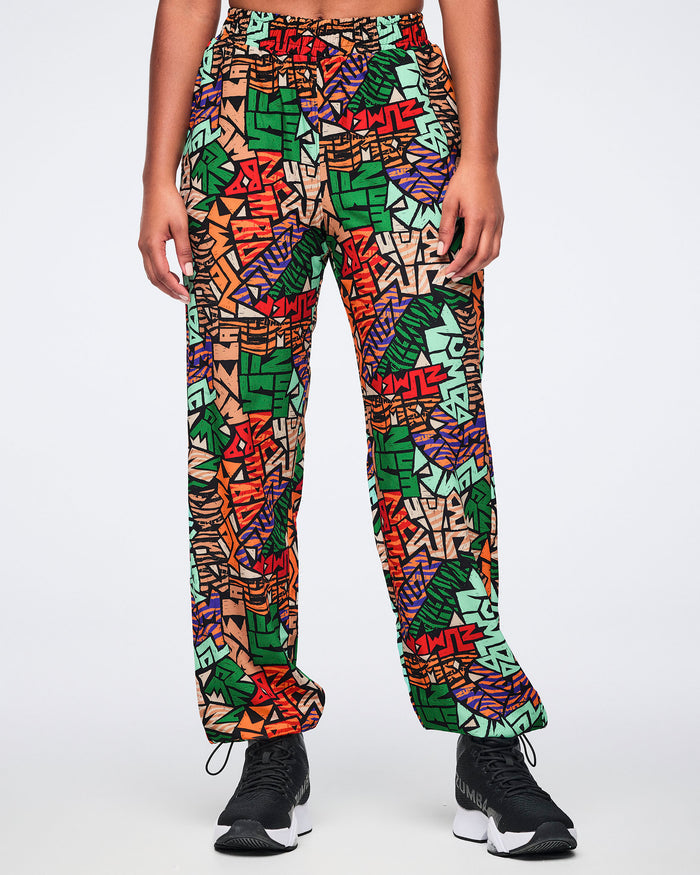 Front view of multicolor Zumba print track pants with an elastic waistband, relaxed straight-leg fit, side pockets, and adjustable ankle bungee cords. Female model wears the pants with black athletic sneakers.