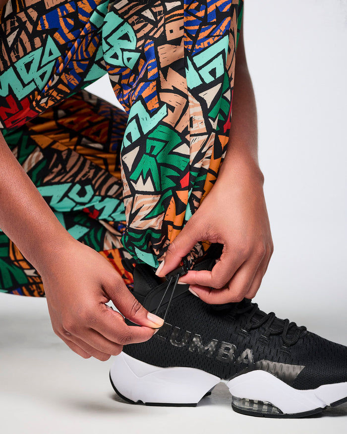 Close-up of multicolor Zumba print track pants showing the adjustable ankle bungee cord at the hem. Female model wears the pants with black Zumba athletic sneakers.