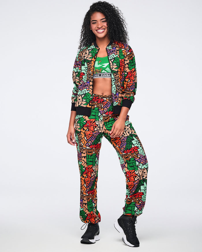 Multicolor Zumba print track pants with an elastic waistband, relaxed fit, side pockets, and gathered ankle cuffs. Female model wears the pants with a matching print jacket, green sports bra, and black athletic sneakers.
