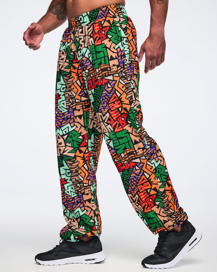 Side view of multicolor Zumba print track pants with an elastic waistband, relaxed fit, and gathered ankle cuffs. Male model wears the pants with black athletic sneakers.