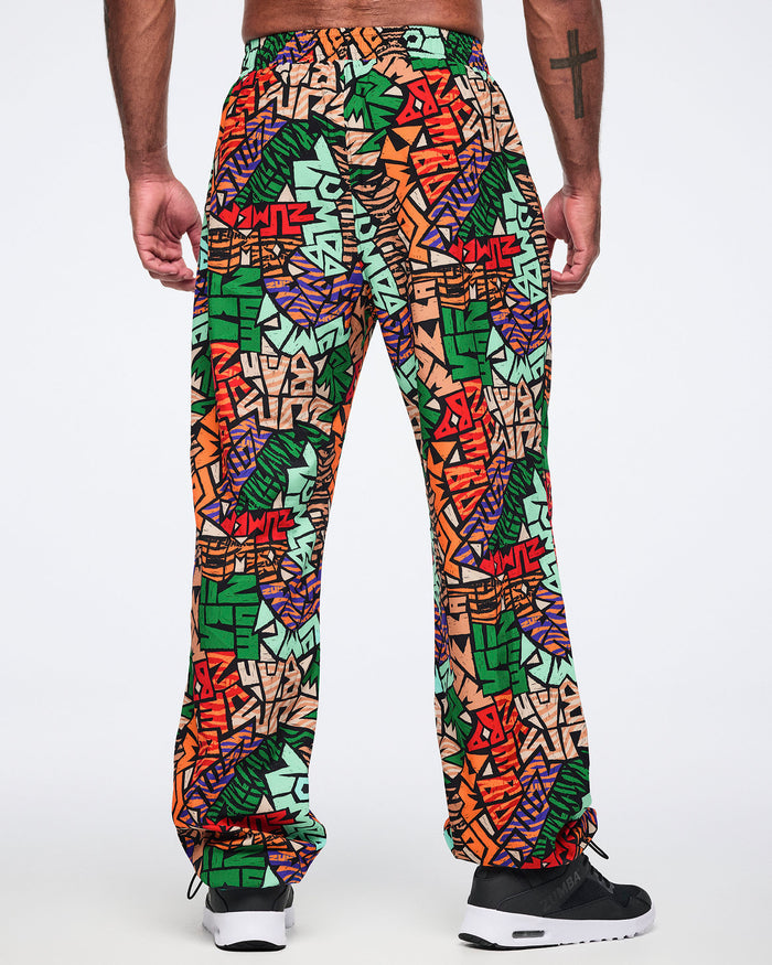 Back view of multicolor Zumba print track pants with an elastic waistband, relaxed fit, and gathered ankle cuffs. Male model wears the pants with black athletic sneakers.