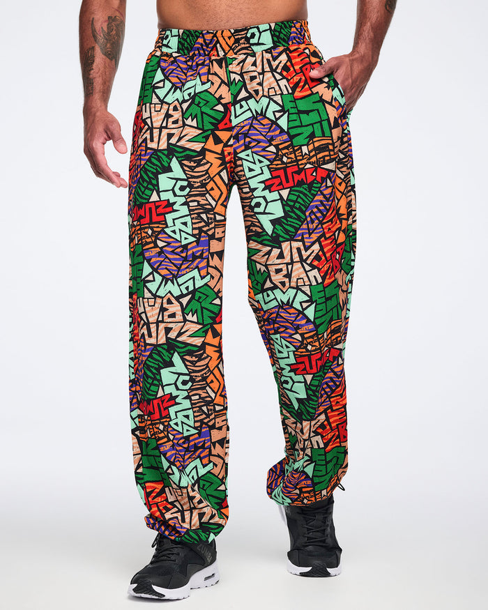Front view of multicolor Zumba print track pants with an elastic waistband, side pockets, relaxed fit, and gathered ankle cuffs. Male model wears the pants with black athletic sneakers.