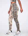 Side view of beige and black mixed-print high-waisted leggings with a fitted full-length silhouette, combining zebra stripes and abstract geometric panels. Female model wears the leggings with white high-top Zumba sneakers and a matching beige and black crop top.