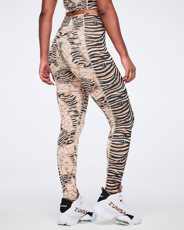 Back view of beige and black mixed-print high-waisted leggings with a fitted full-length silhouette, combining zebra stripes and abstract geometric panels. Female model wears the leggings with white high-top Zumba sneakers and a matching beige and black crop top.