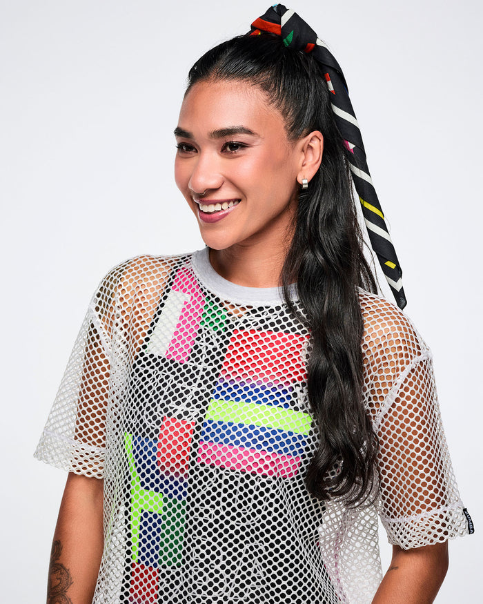 Female model wearing a white mesh top over a multi-colored bodysuit wearing a bandana as a hair tie.