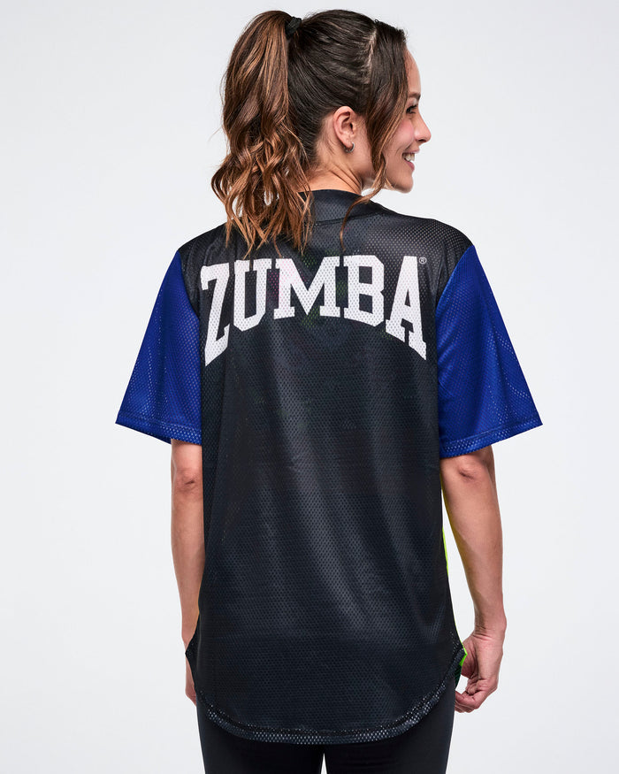Back view of a female model wearing a black Zumba shirt with blue sleeves with "Zumba" in white text, on a white background.