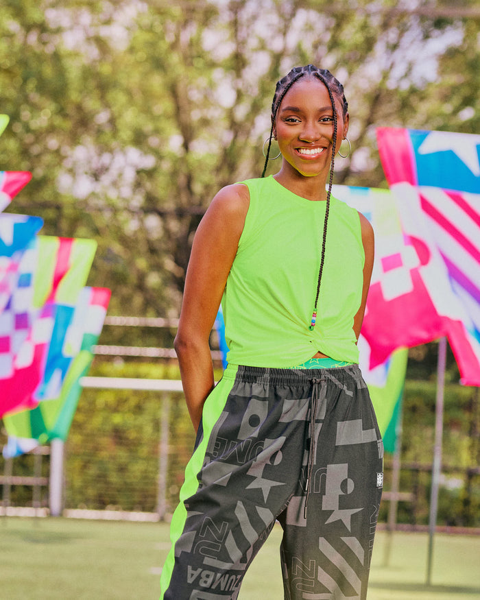Female model outdoors wearing a neon yellow sleeveless top and black pants with patterns, standing in front of colorful flag like patterns.
