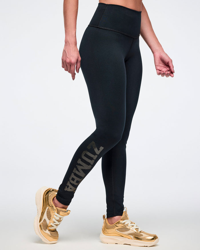 Female model wearing black leggings with gold text and gold sneakers on a white background.