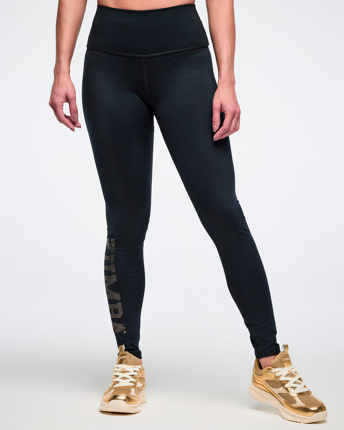 Female model wearing black leggings with gold 'Zumba' text and gold sneakers on a white background.