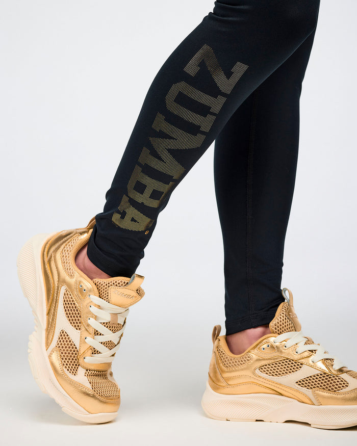 Close-up view of female model right leg wearing black leggings with gold text and gold sneakers on a white background.