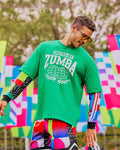 Male model wearing a green 'United by Zumba' t-shirt with colorful shorts in front of a colorful flag outdoor background.